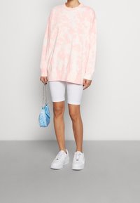 Sudadera de tie-dye en rosa claro, pantalones cortos blancos de ciclismo, zapatillas blancas y un bolso azul con estampado y cadena plateada.