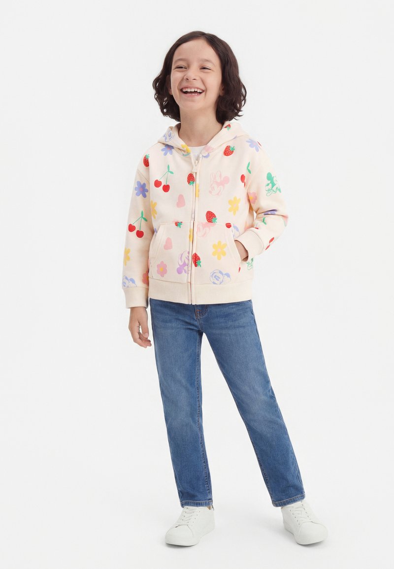 Χαμογελαστό παιδί που στέκεται με το ένα χέρι στην τσέπη, φοράει μια κρεμ φούτερ με φερμουάρ και τυπώματα φρούτων και λουλουδιών, μπλε τζιν και λευκά αθλητικά παπούτσια.