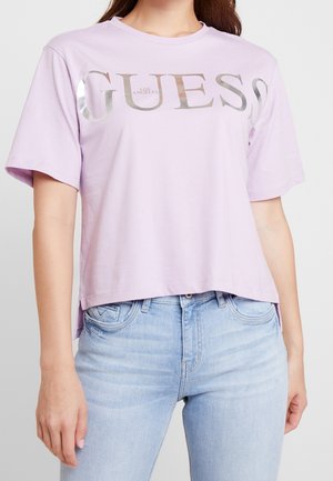 T-shirt en coton violet à manches courtes présentant un logo "GUESS" métallique argenté. Coupe courte au-dessus de la taille avec un col arrondi.