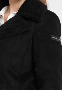 Czarna zamszowa kurtka z dużym, teksturowanym czarnym kołnierzem. Posiada naszywkę z logo na rękawie oraz wyraźnie zarysowaną konstrukcję.