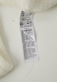 Hvid fleece-stof med etikette, der viser plejeanvisninger og materialekomposition, herunder polyester, polyamid og information om genanvendelse.