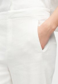 Roka v notranjem žepu belih hlač iz lahkega blaga, ki poudarjajo podrobnosti žepa in pasu.