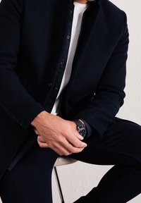 Tmavě modrý kabát z vlněné směsi s vysokým límcem, černými knoflíky a přiléhavým střihem, doplněný o bílé tričko a černé kalhoty. Na zápěstí je viditelný hodinky.