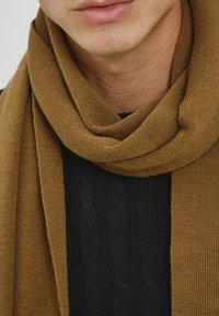 Brown knitted scarf wrapped around the neck, featuring a textured surface, paired with a dark cable-knit sweater underneath.