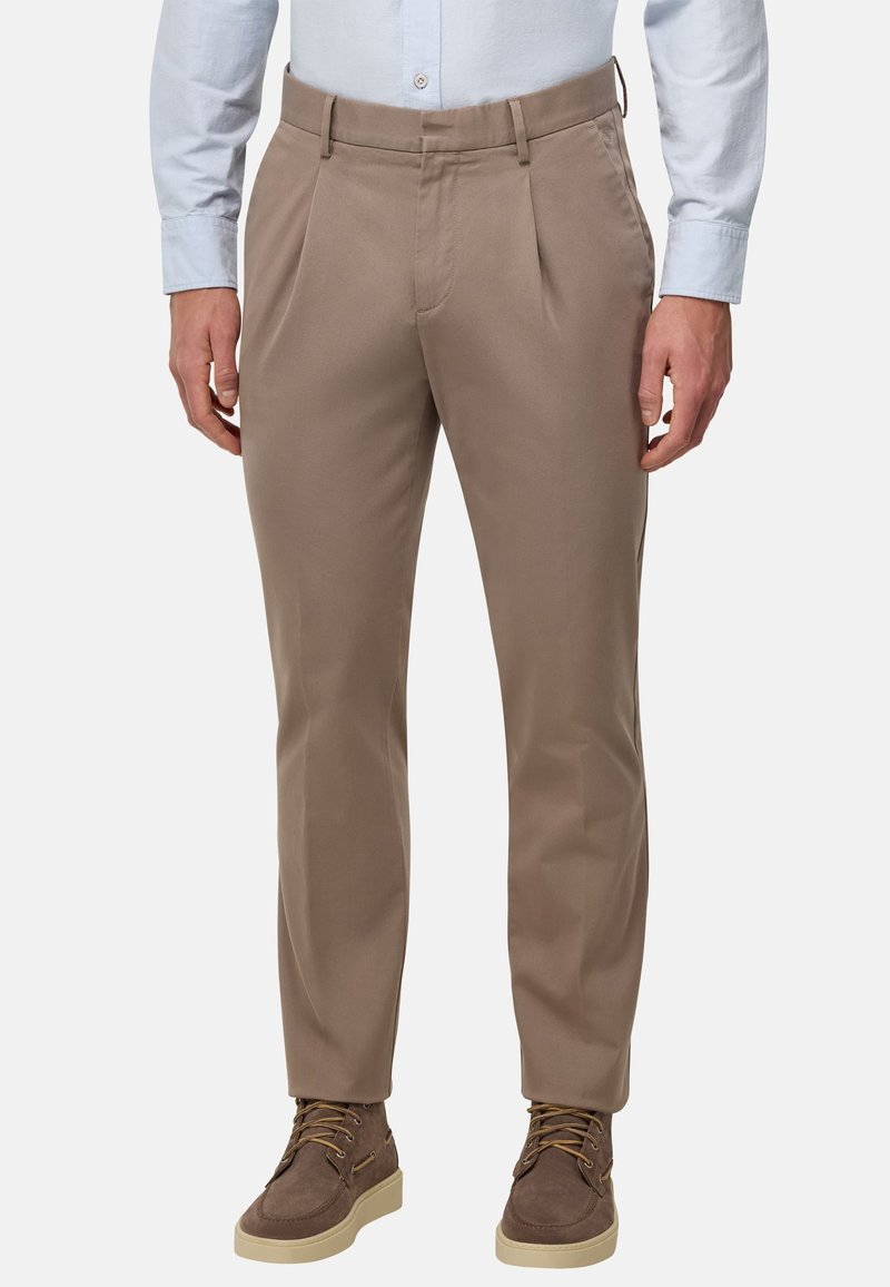 Pantalones beige con pliegues, hechos de un tejido suave, presentan una cintura ajustada. Combinados con zapatos de ante marrones con suelas crema.