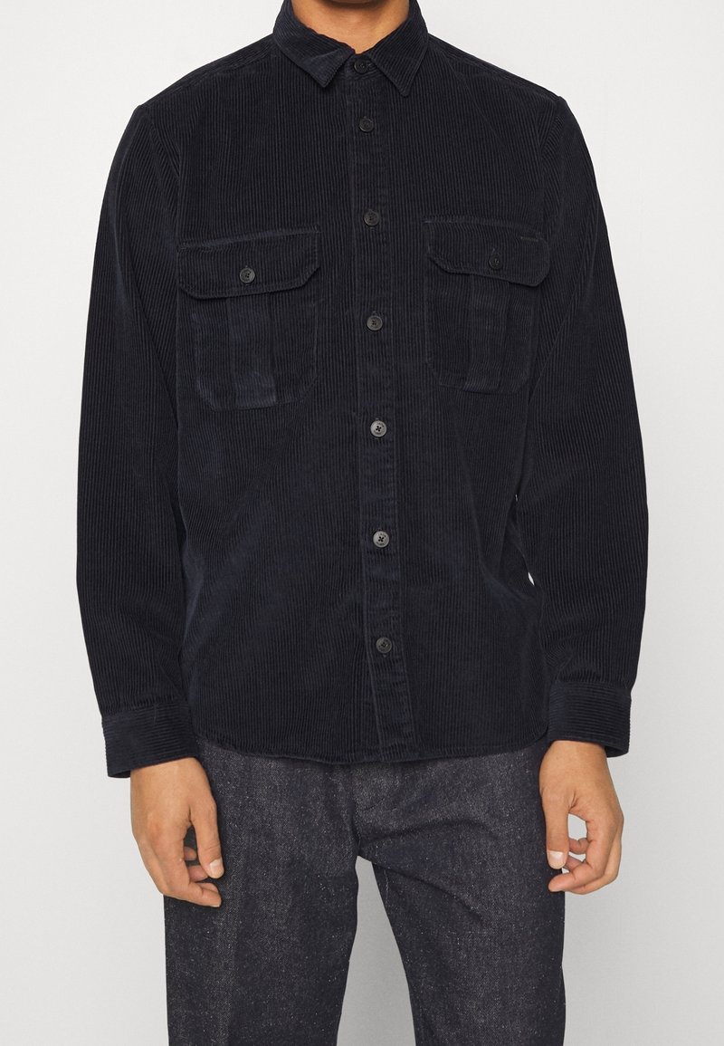 Mand iført sort fløjlsribbede skjorte med knapper og to brystlommer samt mørke bukser foran en ensfarvet baggrund.