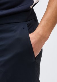 Άτομο που φοράει μπλε παντελόνια με το χέρι εν μέρει στην πλαϊνή τσέπη, εμφανίζοντας ομαλό ύφασμα και μικρό κεντημένο λογότυπο κοντά στην τσέπη.