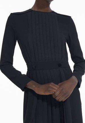 Nainen pukeutuneena pitkähihaiseen mustaan mekkoon, jossa on pystysuoria laskoksia yläosassa ja kaksi nappia vyötäröllä, kädet yhdessä edessä.