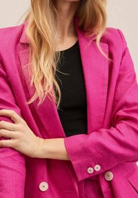 Femme blonde aux cheveux longs portant un blazer rose vif à double boutonnage sur un haut noir, les bras croisés sur le torse.