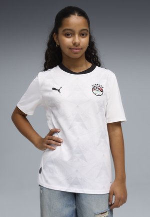 Jeune personne aux cheveux bouclés portant un maillot de football blanc Puma Égypte et un jean bleu clair déchiré, debout avec une main sur la hanche.