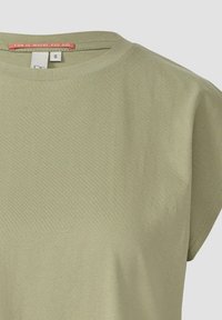 Lichtgroene T-shirt met korte mouwen van zachte stof. Heeft een ronde hals en een label met de tekst "PLEZIER IS WAAR JIJ BENT" op de kraag.