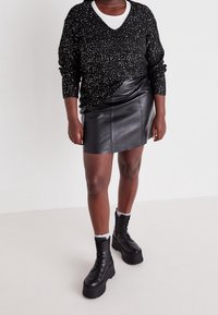 Pull noir à motifs tachetés, col en V, manches longues ; mini jupe en cuir noir ; bottes à lacets noires à plateforme avec semelles épaisses ; sous-vêtement blanc.
