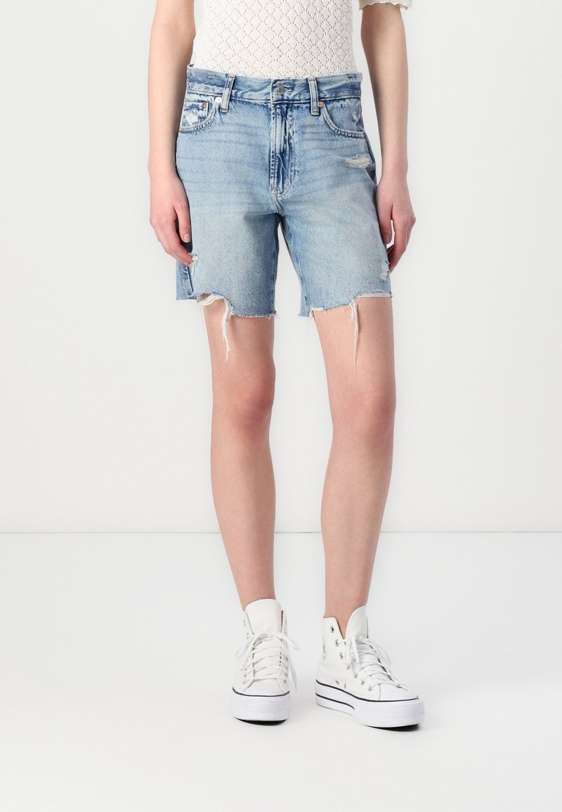 Personne portant un short en jean délavé bleu clair avec des ourlets effilochés, des baskets montantes blanches et un haut blanc texturé à manches courtes.
