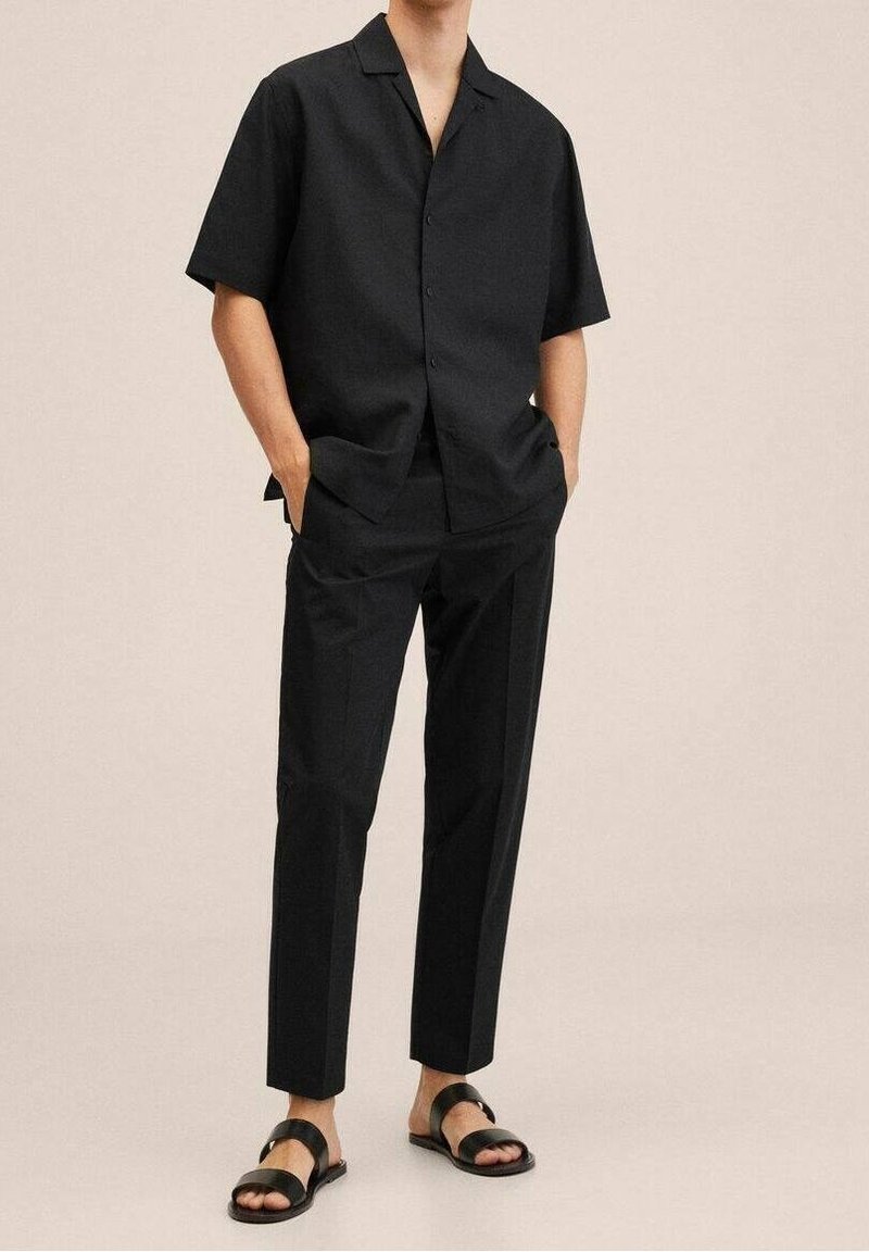 Homme portant une chemise noire à manches courtes boutonnée, un pantalon noir sur mesure et des sandales noires à deux brides, mains dans les poches, fond beige.