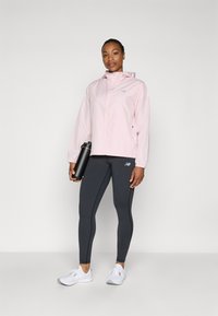 Light pink zip-up jacket with hood, black leggings featuring side accents, and white athletic shoes. Black water bottle held in hand.