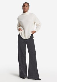 Weißer Strickpullover mit Rollkragen, gerippten Bündchen und Saum, kombiniert mit dunkelblauen Jeans mit weitem Bein. Das Model steht mit den Händen in den Taschen und trägt schwarze Pumps.