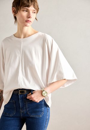 Løssete, lys rosa T-skjorte med rund hals og senkede skuldre, sammen med mørkeblå jeans og et lysegrønt armbåndsur.