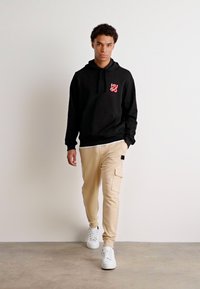 Svart huvtröja med röd logotyp, ihop med beige cargobyxor med sidofickor och uppvikta anklar. Vita sneakers fulländar looken.