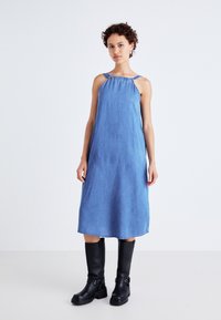 Denim midi-kjole med avslappet passform, justerbare smale stropper og rynket utringning, kombinert med knelengde svarte støvler.