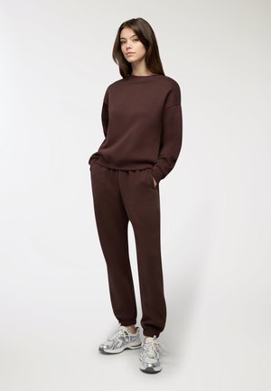 Felpa marrone e pantaloni jogger abbinati, entrambi in un tessuto morbido. L'outfit presenta una vestibilità rilassata, polsini elastici e tasche laterali. Sneakers argentate completano il look.