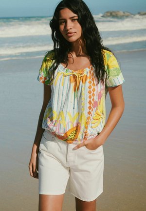 Femeie cu păr lung și închis la culoare, purtând o bluză colorată cu model și pantaloni scurți albi, stând pe o plajă lângă ocean.