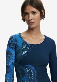 Langärmliges Oberteil in Blau mit Muster, weitem Rundhalsausschnitt und strukturiertem Rippstoff. Es zeigt florale und abstrakte Designs in verschiedenen Blautönen.