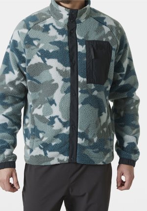 Man draagt een blauw, grijs en wit camouflagefleecejack met zwarte drukknopsluiting en ritszak op de borst, gecombineerd met een zwarte broek.
