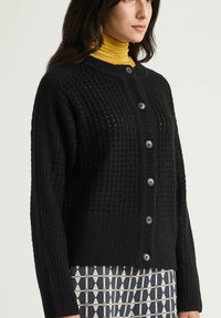 Cardigan noir en maille avec un motif texturé, boutonné à l'avant et poignets côtelés. Porté sur un col roulé moutarde, complété par un pantalon à motifs.