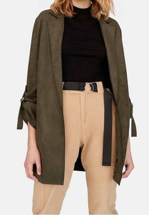 Kobieta ubrana w oliwkowo-zielony, długi blezer, czarną bluzkę oraz beżowe spodnie z wysokim stanem i czarnym paskiem z metalową, srebrną klamrą w kształcie pierścienia.