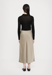 Svart langermet topp i gjennomsiktig tekstur, sammen med et beige satengskjørt som faller til midten av leggen. Modellen har på seg enkle svarte slip-on sko.