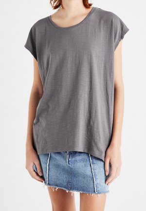 Femme portant un haut ample à manches courtes gris et une mini-jupe en denim bleu clair effilochée, debout sur un fond blanc.