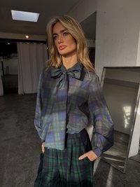 Sheer plaid blouse in green and blue with a large bow tie at the collar, paired with dark green and black pleated trousers.