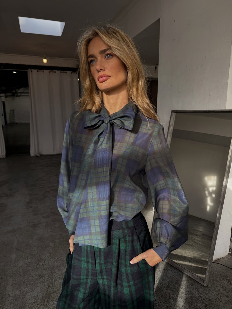 Sheer plaid blouse in green and blue with a large bow tie at the collar, paired with dark green and black pleated trousers.