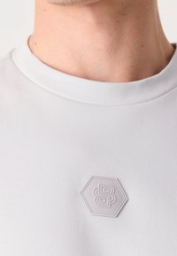 Personne portant un t-shirt blanc à col rond avec un patch logo hexagonal en relief sur la poitrine, présentant des lettres entrelacées.