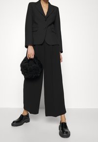 Woman wearing black wide-leg pants, black blazer, black chunky loafers, holding a small black fuzzy handbag with gold rings on left hand.