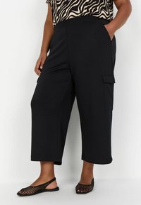 Pantalons cargo noirs raccourcis avec poches latérales, fabriqués en tissu doux. Associés à des ballerines à dos en maille noires et un haut à motifs.