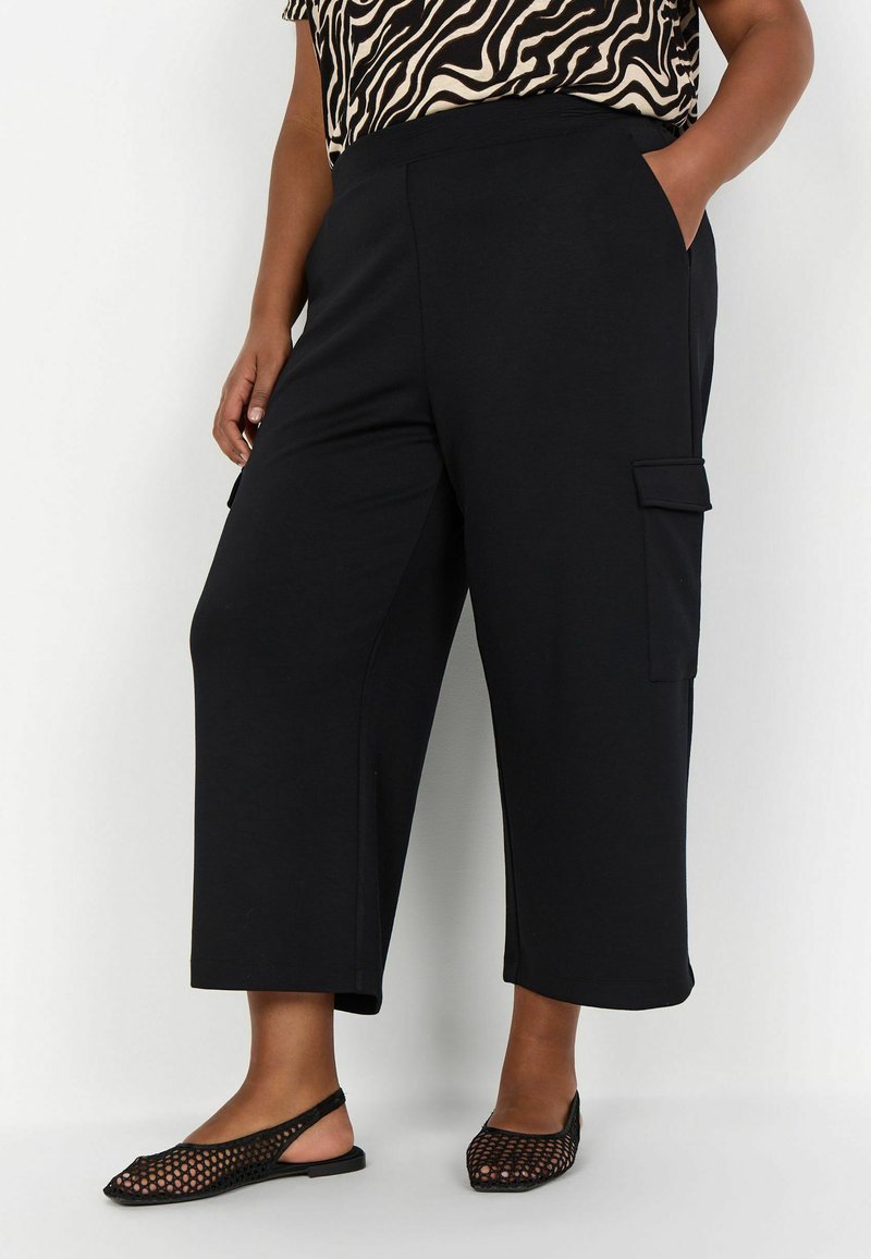 Pantalons cargo noirs raccourcis avec poches latérales, fabriqués en tissu doux. Associés à des ballerines à dos en maille noires et un haut à motifs.