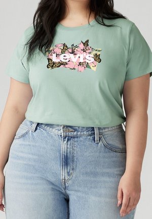 Nainen pukeutuneena vaaleanvihreään Levi's-t-paitaan, jossa on pinkkejä kukkia ja keltaisia perhosia, yhdistettynä vaaleansinisiin korkeavyötäröisiin farkkuihin.