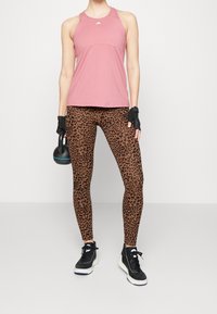 Canottiera sportiva rosa chiaro con schiena a incrocio, abbinata a leggins marroni con stampa leopardata nera. Scarpe da allenamento nere e un kettlebell.