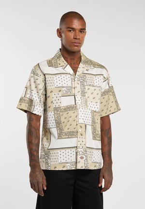 Camisa de manga corta en beige y blanco con patrones variados, incluyendo diseños de paisley y geométricos, confeccionada con tela ligera.