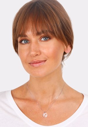 Silberne Halskette mit einem runden Anhänger mit ausgeschnittenem Herz-Detail, getragen von einem Modell mit einem weißen Oberteil und neutralem Hintergrund.