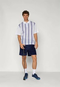Hvit kortermet polo med blå vertikale striper, kombinert med marineblå shorts og blå joggesko. Står med en golfkølle.