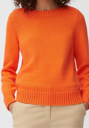 Person trägt einen leuchtend orangefarbenen Strickpullover mit geripptem Saum und Bündchen, kombiniert mit beigen Hosen, Hände in den Taschen, neutraler Hintergrund.