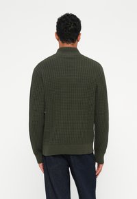 Pulover texturat de culoare verde închis, cu guler înalt și mansete cu dungi, având un model de tricotaj în stil vafe și un tiv drept.