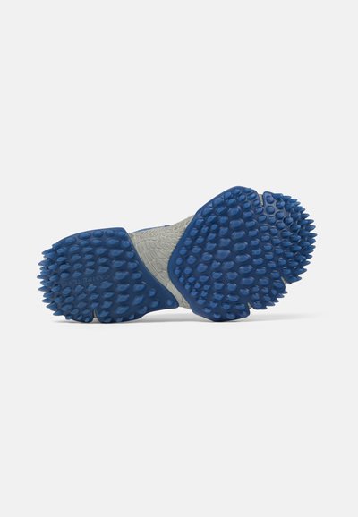 Suela de un zapato con tacos de goma azul en forma de vaina que cubren las zonas del talón y la parte delantera del pie sobre un fondo gris claro.