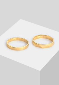 Deux bagues en or présentées sur une surface géométrique ; l'une est lisse et ronde, l'autre est facettée avec une forme octogonale, toutes deux brillantes.