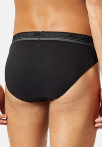 Ein Mann steht vor einem schlichten Hintergrund, mit schwarzer Unterwäsche und einem markierten Bund, der seinen unteren Rücken und die Oberschenkel zeigt.