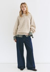 Béžový útulný pletený sveter s raglánovými rukávmi, oblečený na zelenej kárované košeli, skombinovaný s voľnými tmavomodrými džínami a čiernymi topánkami.