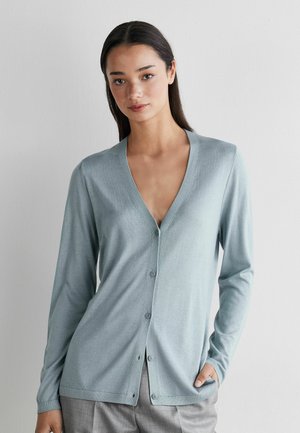 Kvinde iført en lyseblå cardigan med knapper, V-hals og lange ærmer, stående foran en ensfarvet lys grå væg.