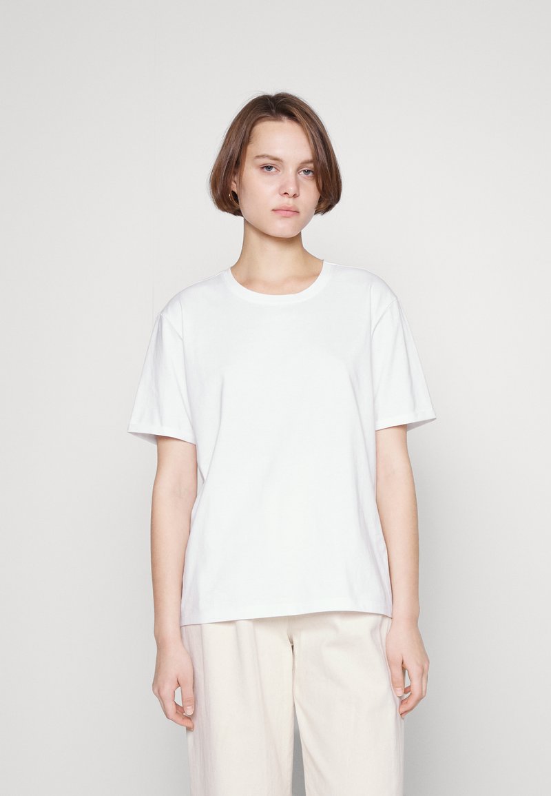 White cotton t-shirt with short sleeves and a round neckline, featuring a relaxed fit and smooth texture. Paired with light-colored trousers.